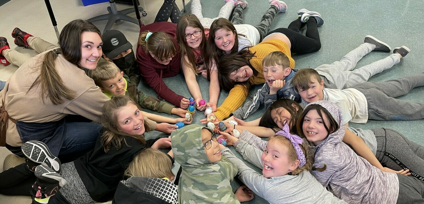 a group of students and their teacher laying on the classroom floor in a circle smiling at the camera